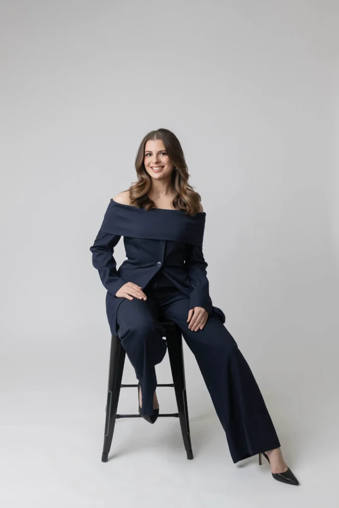 Portrait of a smiling woman in a navy off shoulder suit sitting on a black stool posing with one leg crossed