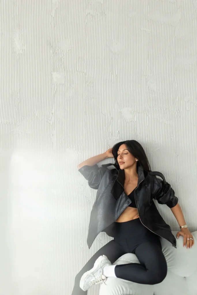 Woman in a black leather jacket and dark outfit sitting on a gray sofa eyes closed against a white textured wall