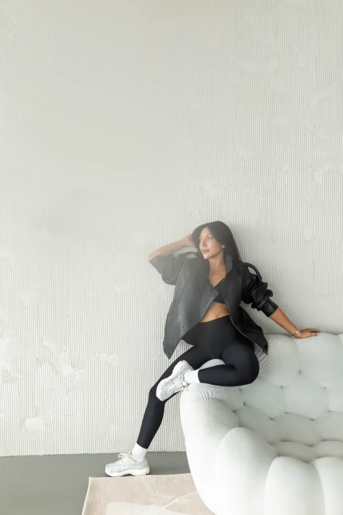 Woman with dark hair sits sideways on a light gray tufted sofa wearing a black outfit and gray leather jacket posing against a textured wall