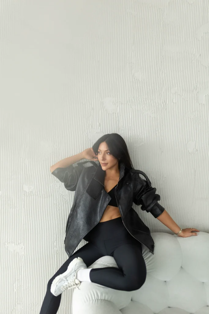 Woman in a black leather jacket and leggings sits on a light gray tufted couch posing with one hand behind her head