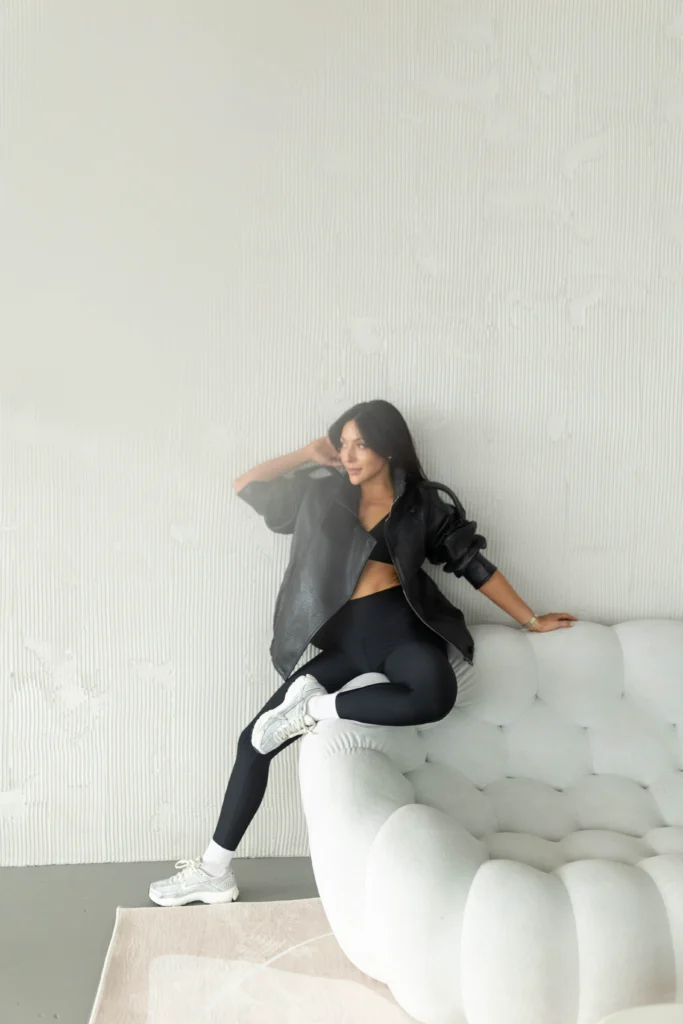 Woman in a black outfit and white sneakers lounging on a light gray tufted sofa against a pale textured wall one arm resting on the back