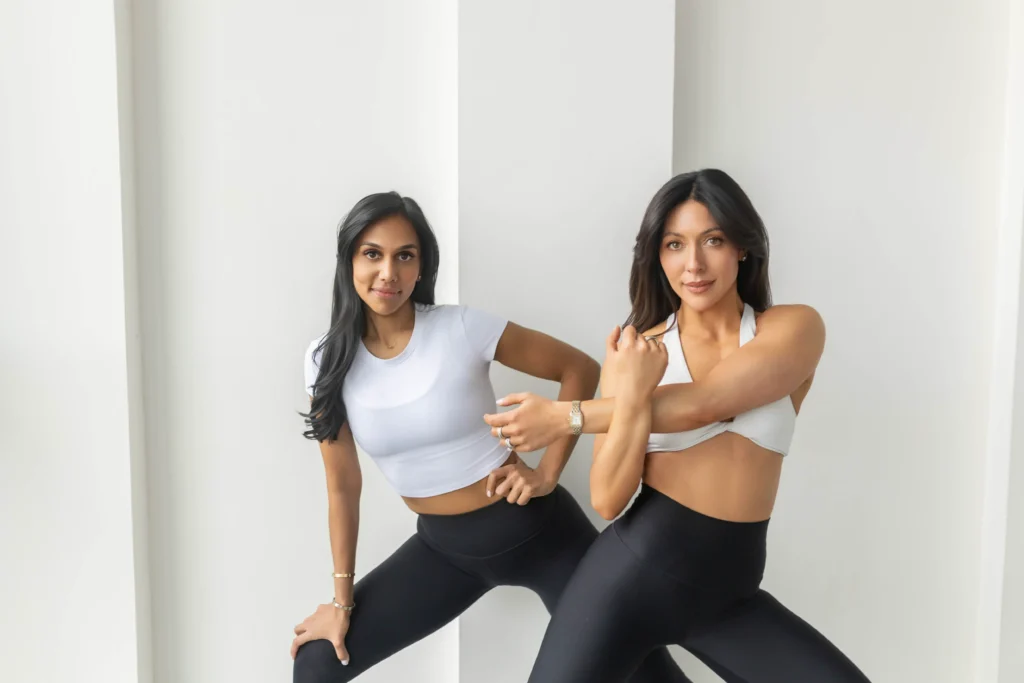 Two women in white athletic tops and black leggings perform a synchronized stretches in a bright minimalist studio setting