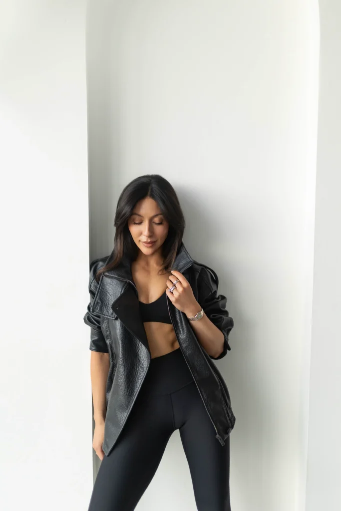 Woman in a black leather jacket and black outfit standing against a pale wall looking down at her hand