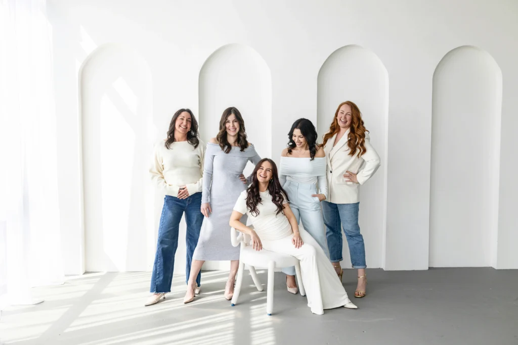 Five women posing together in a bright white studio with tall arched wall panels one seated in a white dress smiling with the group