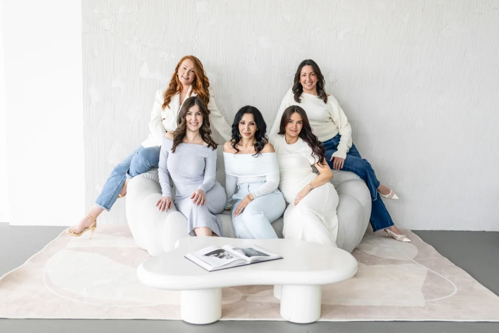 Five women pose together on a white sofa smiling in a bright modern room with a textured wall behind them