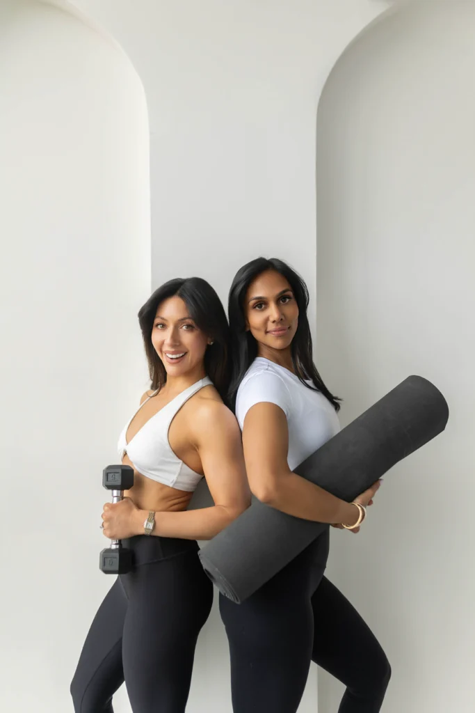 Two women in white tops pose back to back holding a dumbbell and a rolled yoga mat in a bright minimal gym or studio setting
