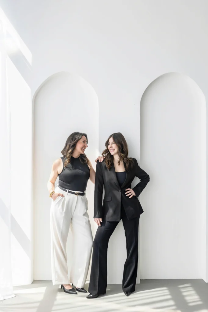 Two women in black and white business attire posing and smiling in a bright white hallway with arched alcoves