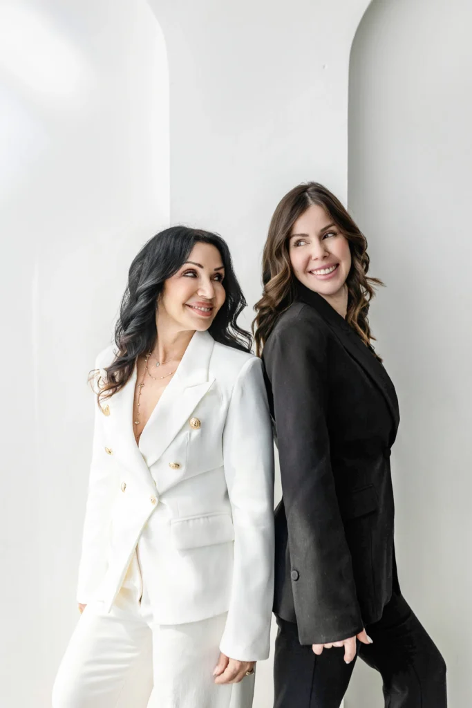 Two professional women in white and black suits standing back to back smiling at each other against alight curved white background
