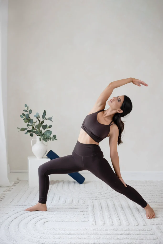 Woman in dark sportswear performs a side stretch yoga pose in a bright minimal room with a plant on a white table nearby