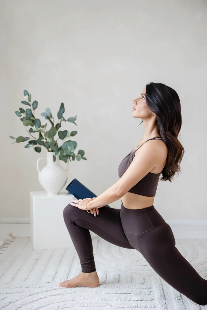 Woman in brown athletic wear performs a lunge in a bright room beside a white vase with eucalyptus branches