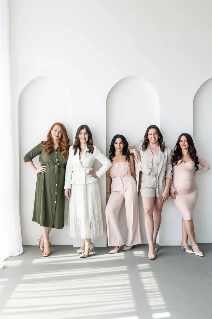 Five women posing together in a bright studio with white arches wearing pastel outfits and heels