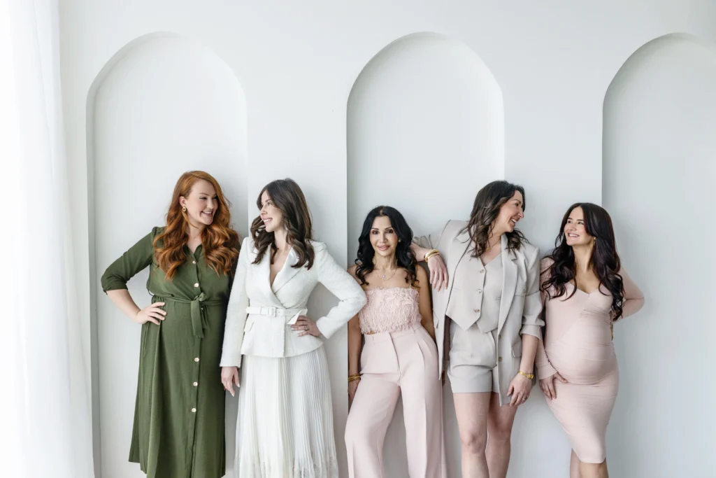 Six women in chic outfits posing together in a bright minimal studio backdrop smiling and looking at each other