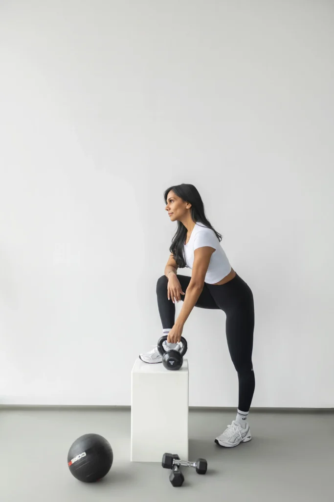 Woman performing a squat with a kettlebell in one hand while standing on a white block in a bright studio gym mats and weights nearby