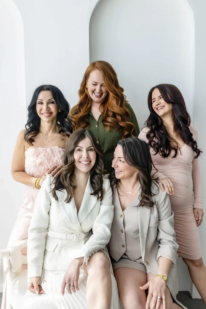 Five women posing together smiling in a bright neutral toned room they wear light pink beige and olive outfits and sit or stand close to one another