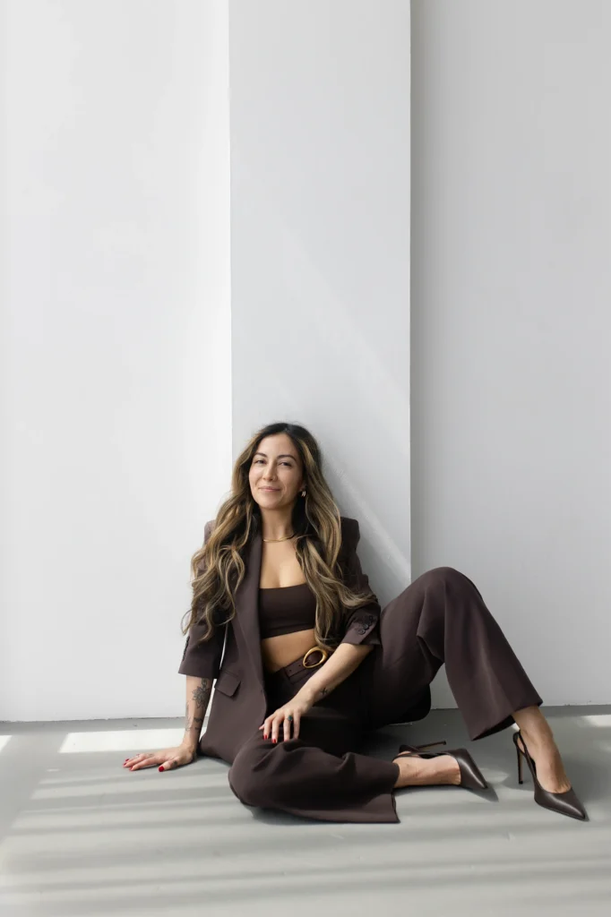 Woman in a brown suit with a crop top seated on the floor against a white wall and looking at the camera