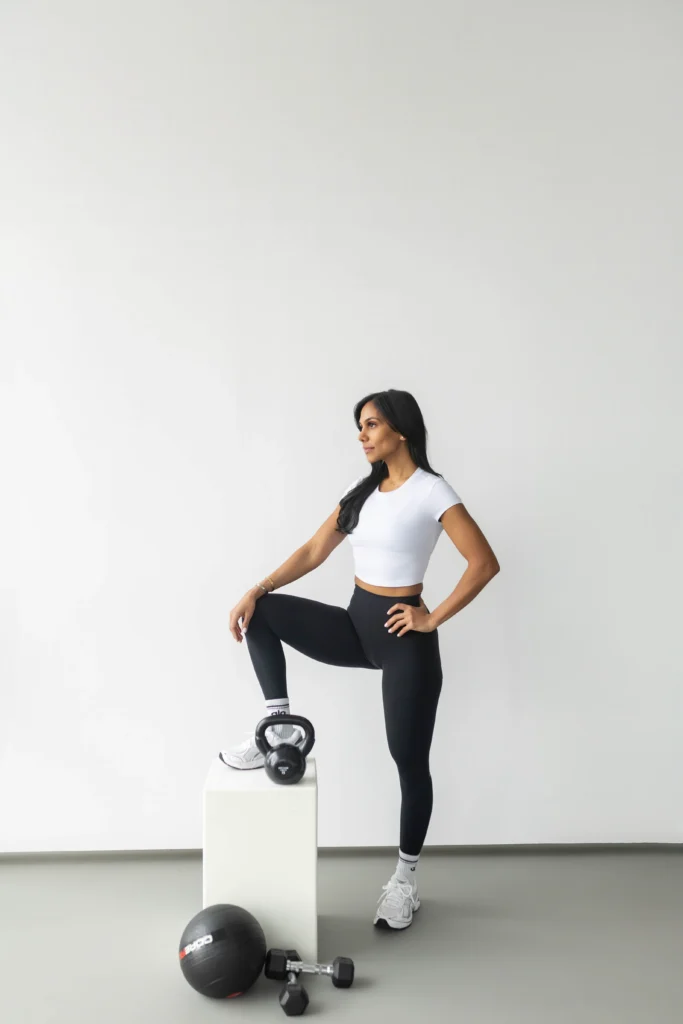 Fit woman in a white crop top and black leggings stands with one foot on a white block hands on hips next to kettlebell dumbbells and a medicine ball on the floor