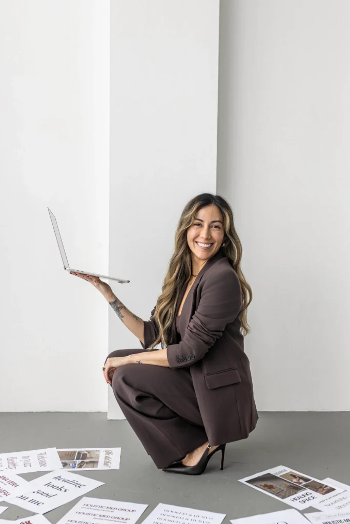 A smiling woman in a brown suit kneels on the floor holding an open laptop outward as if presenting it with scattered papers around her on the gray floor against white walls