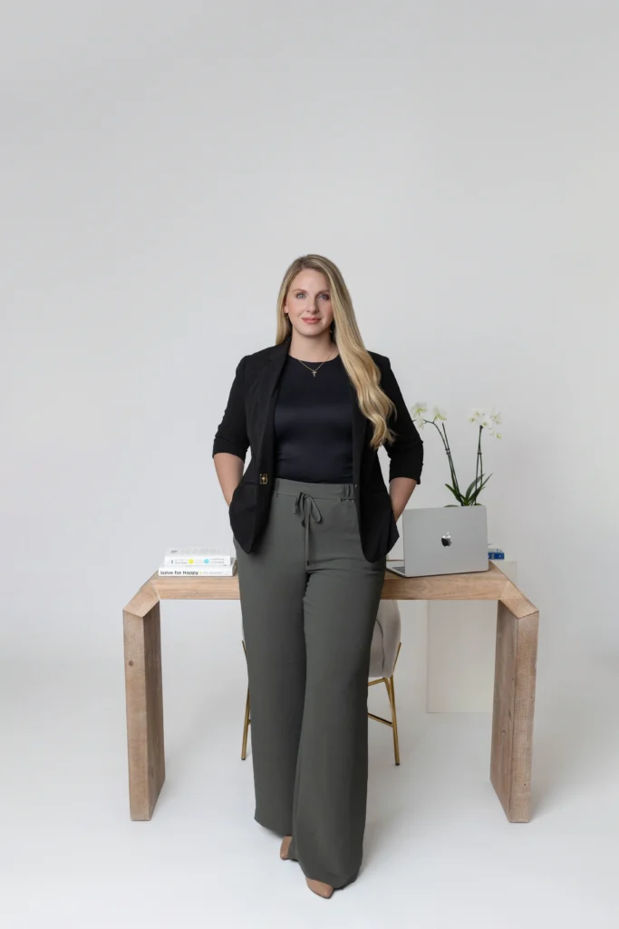 Professional woman stands beside a wooden desk with a laptop and books in a minimalist office setting hands in pockets in a black blazer and dark pants