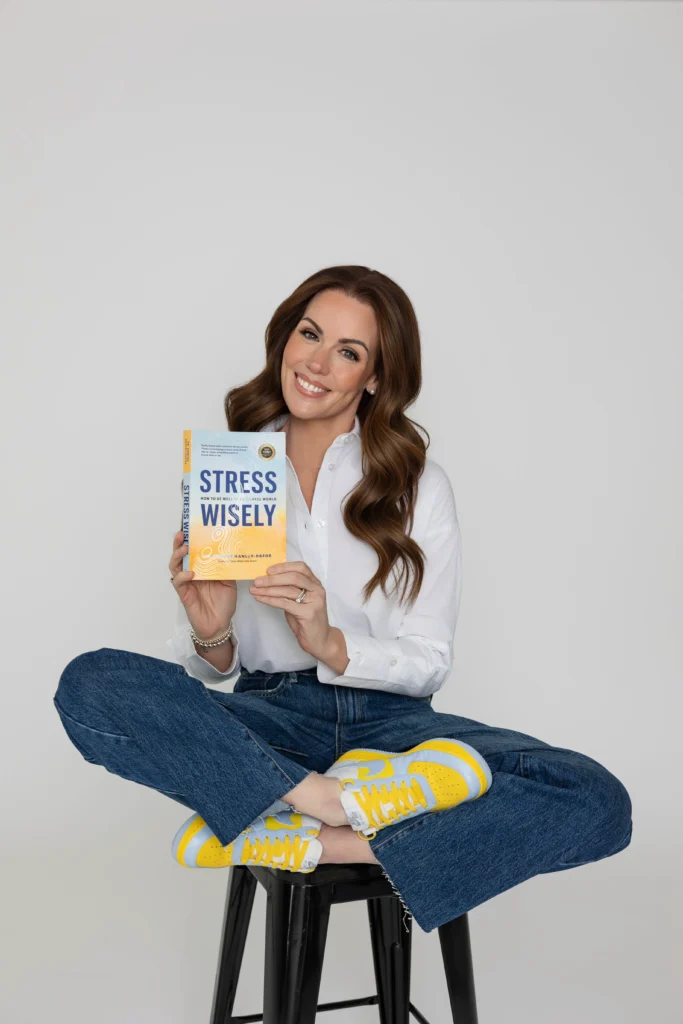 Smiling woman in a white shirt and jeans sits crosslegged on a black stool holding a book titled Stress Wisely