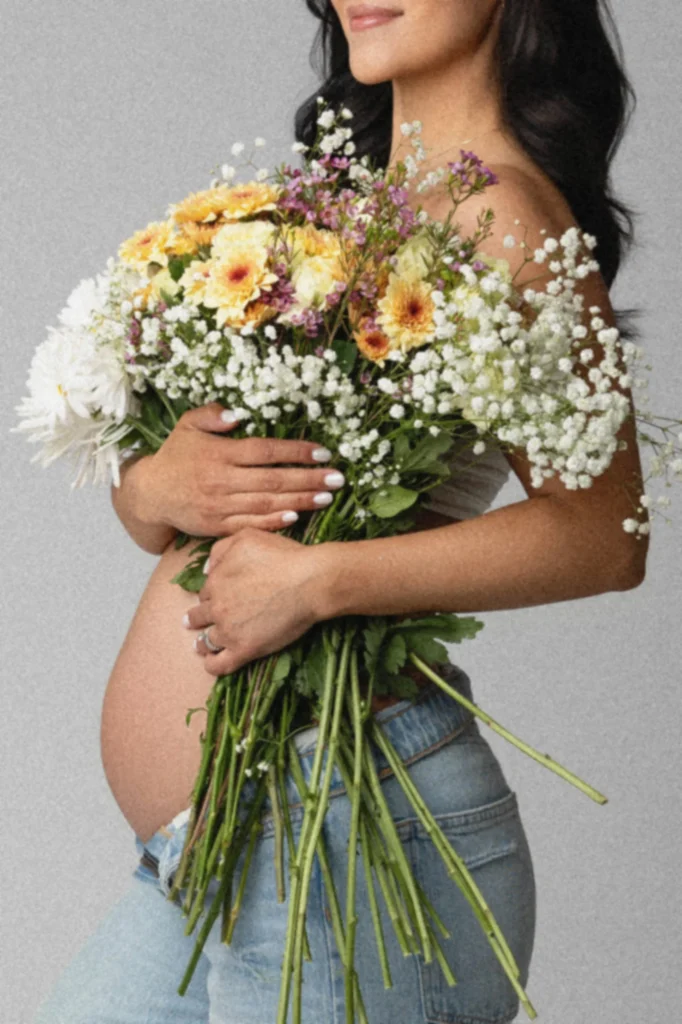 Pregnant woman wearing jeans holds a large bouquet of white babys breath and yellow flowers against a gray backdrop