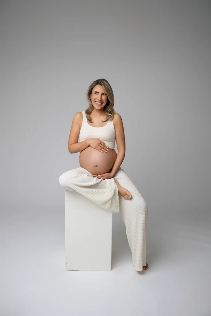 Pregnant woman sits on a white block cradling her belly and smiling in a white outfit against a gray studio background