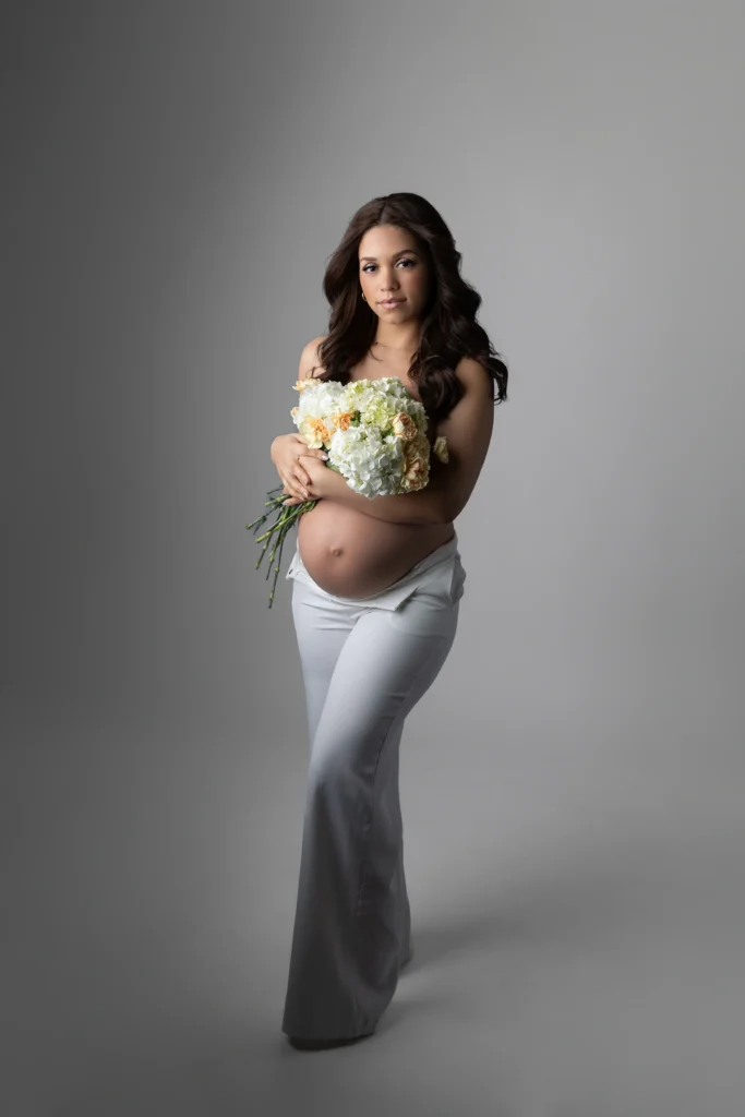Pregnant woman with dark hair wearing white pants cradling a bouquet of white and peach flowers against a gray background