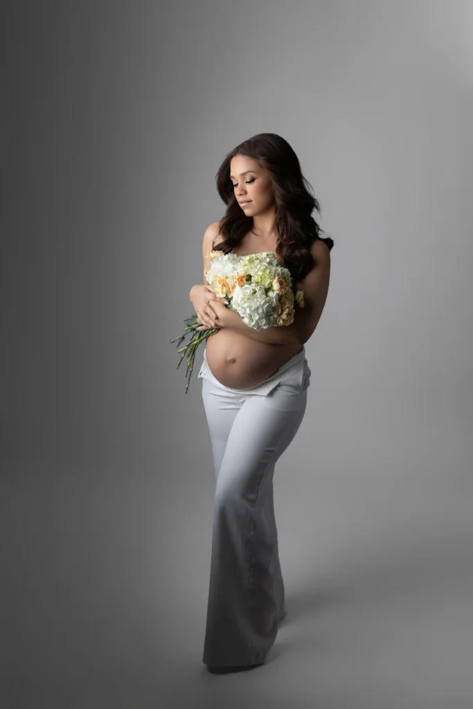 Pregnant woman in white pants cradling a bouquet of white flowers looking down thoughtfully against a gray backdrop
