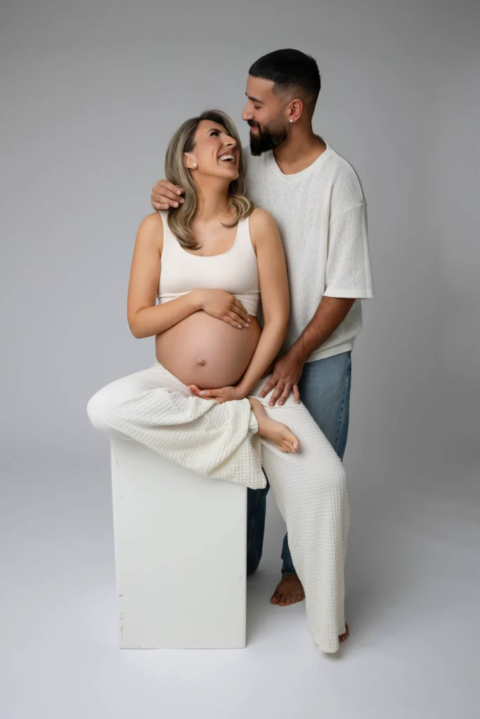 Pregnant woman seated on a white block smiling as her partner stands behind her with a hand on her shoulder