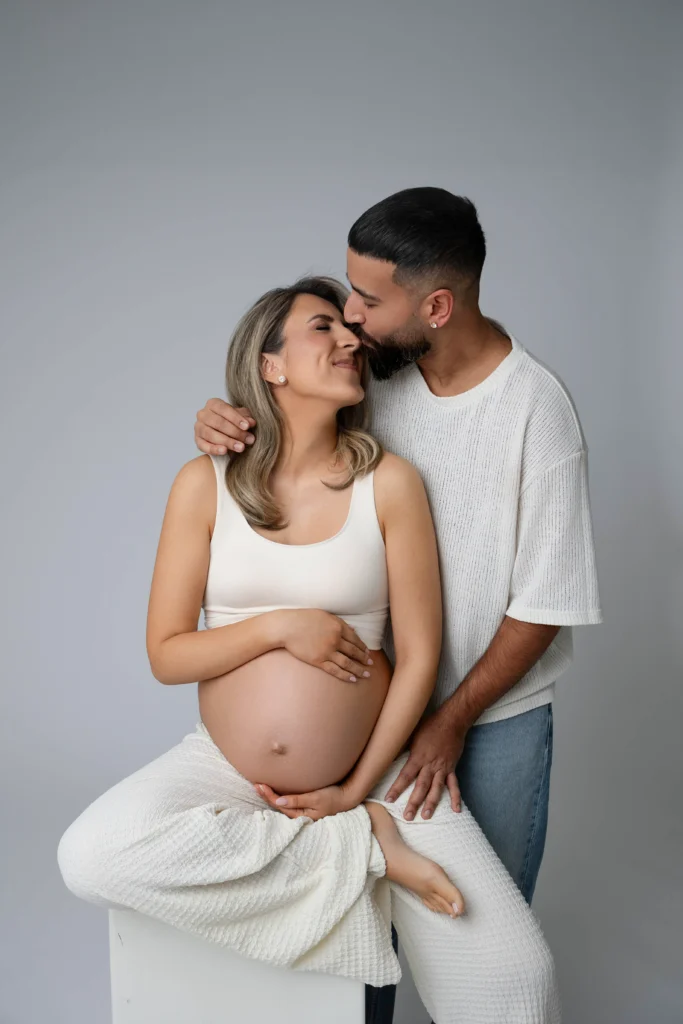 Pregnant woman in a white top sits while her partner hugs her from behind and kisses her on the head or temple cradling her belly with her hands