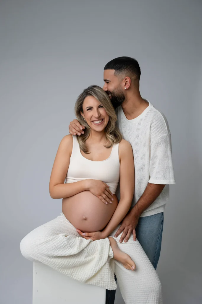 Pregnant woman smiling while seated with her partner standing behind and wrapping an arm around her shoulders