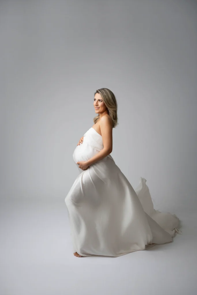 Pregnant woman in a flowing white gown side view cradling her belly in a studio with a gray backdrop