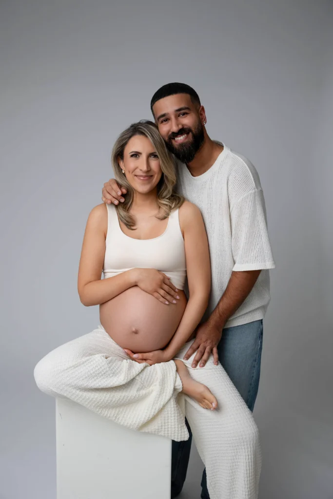 Pregnant woman sitting on a white block with a partner standing behind her his arm around her shoulders as they smile