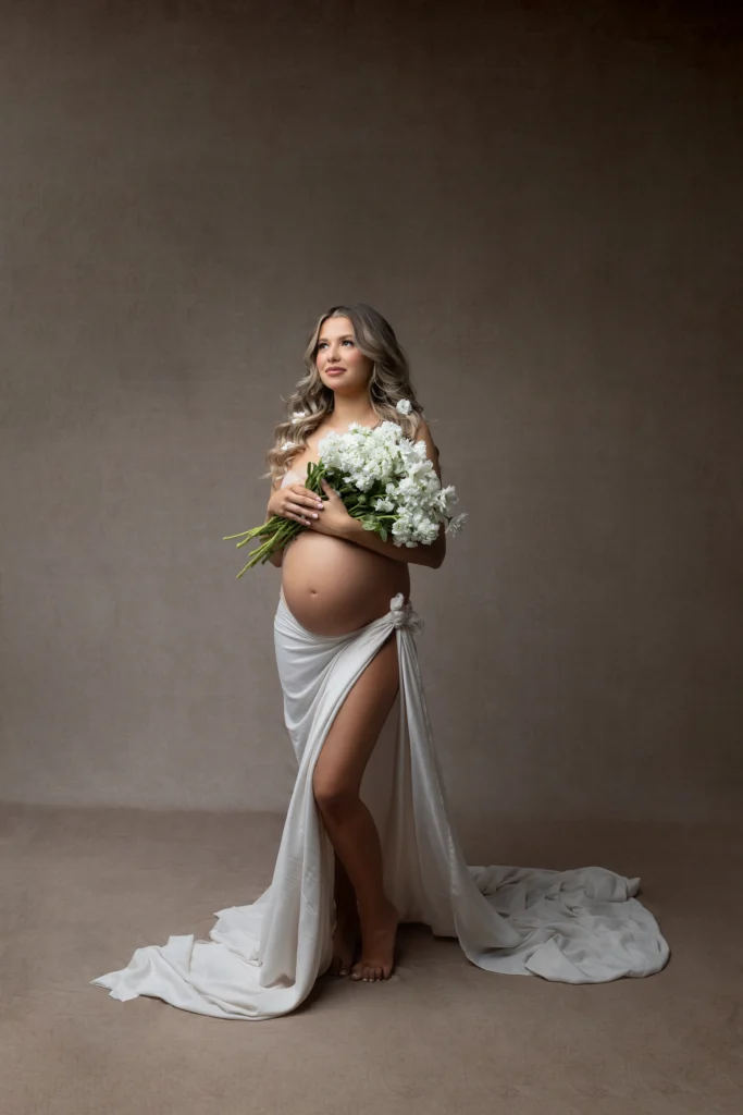 Pregnant woman standing in a neutral studio wearing a white wrap dress and holding a bouquet of white flowers
