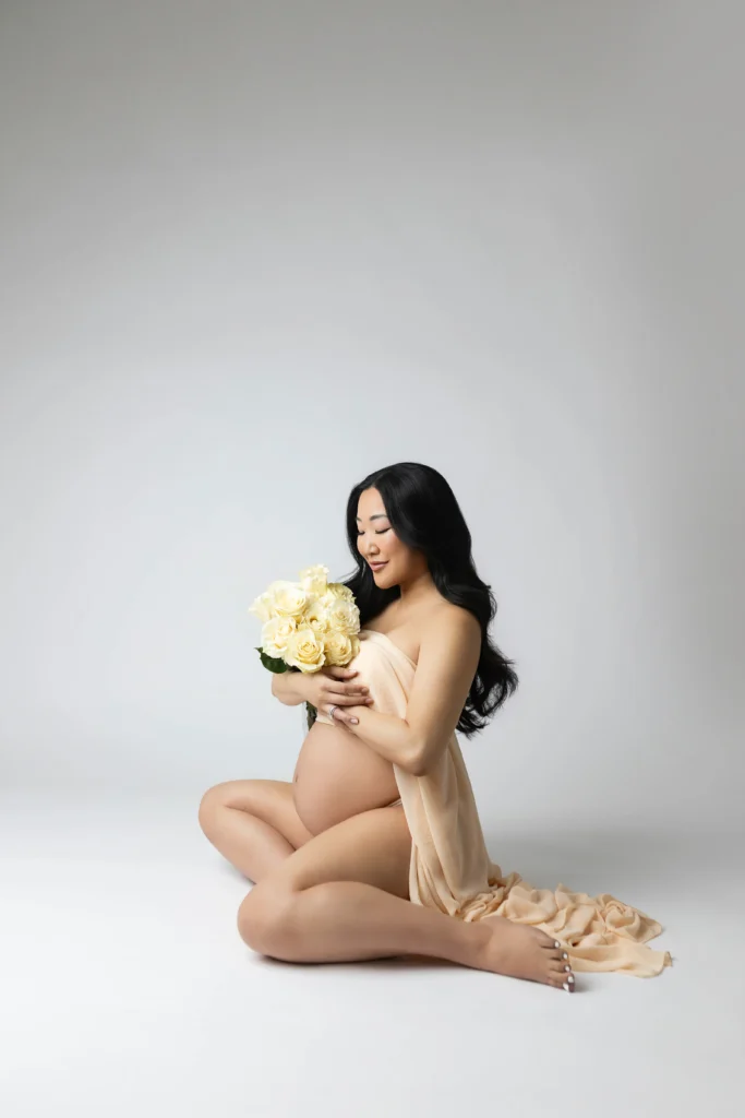 Pregnant woman kneeling on a white studio floor cradling a bouquet of white roses with a soft beige wrap