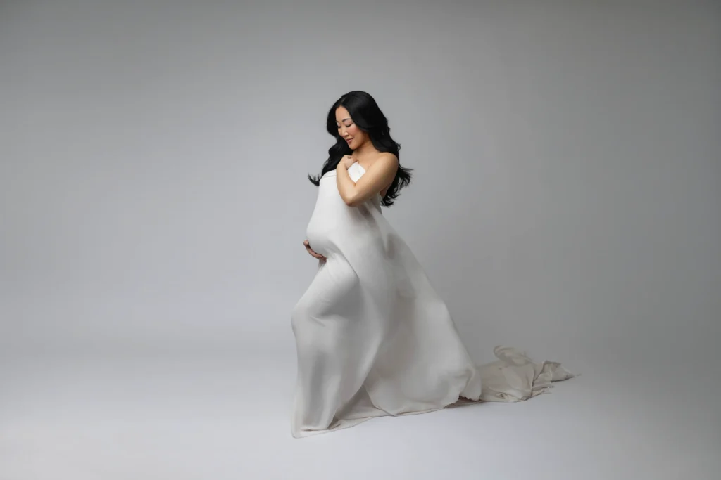 Pregnant woman in a white flowing dress gently cradling her belly against a neutral gray backdrop