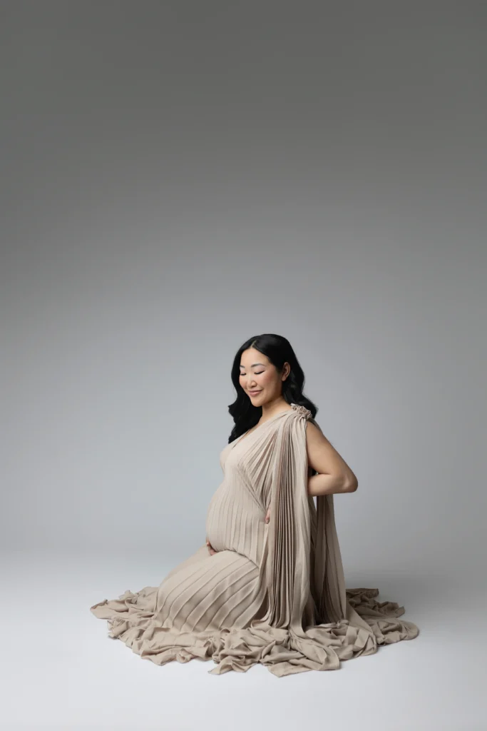 Pregnant woman in a beige pleated gown sitting on a light gray studio floor in a posed portrait
