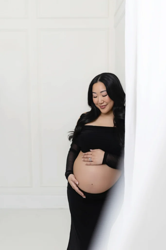 Pregnant woman in a black off the shoulder dress standing beside a white curtain cradling her belly with a gentle smile