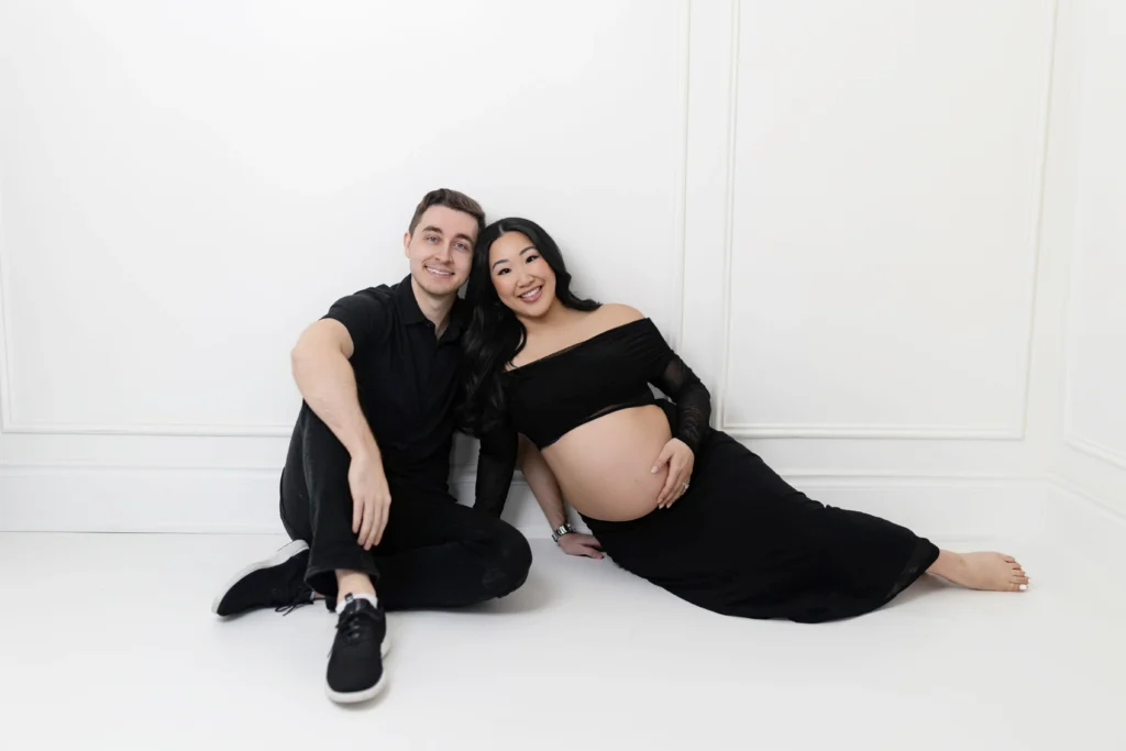 Smiling couple sitting on the floor against a white wall pregnant woman in a black dress rests a hand on her baby bump beside her partner in black