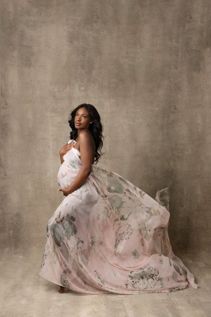 Pregnant woman in a pastel floral maternity gown stands against a textured beige studio wall