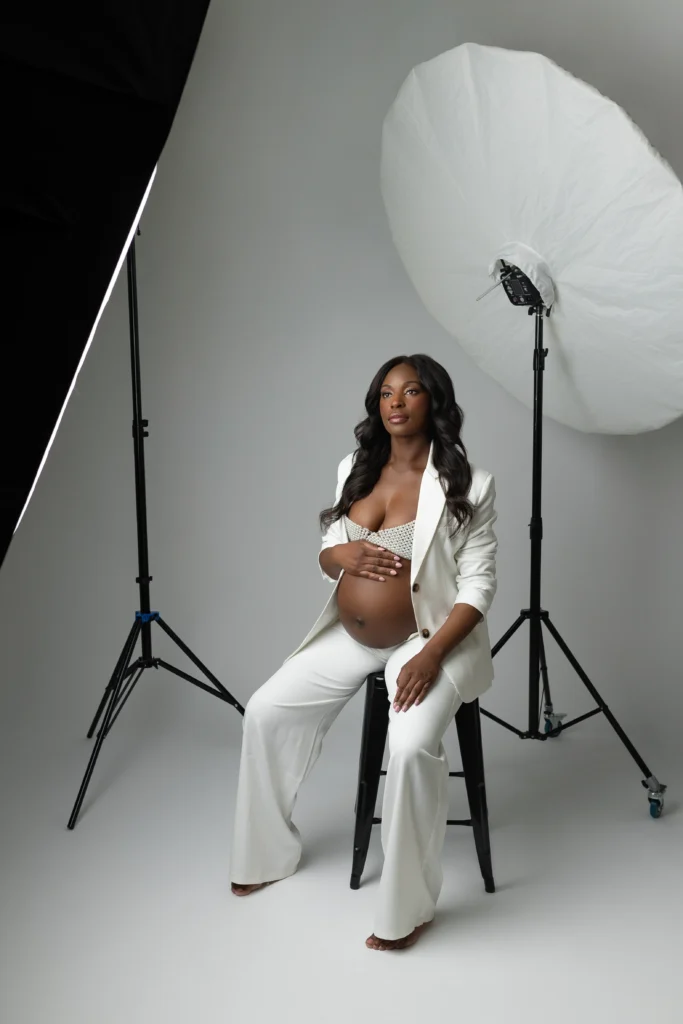 Pregnant woman in a white suit sits on a stool in a photography studio cradling her baby bump