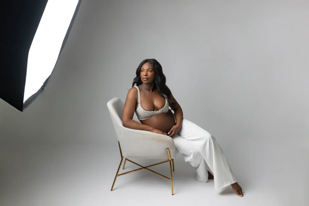 Pregnant model posing in a studio chair wearing a beaded white top and white pants with a large studio light in the background