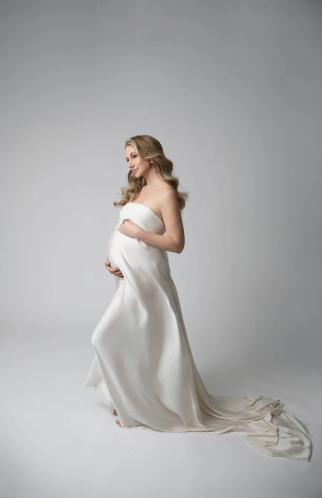 Pregnant woman in a flowing white gown posing in profile against a gray studio backdrop cradling her belly