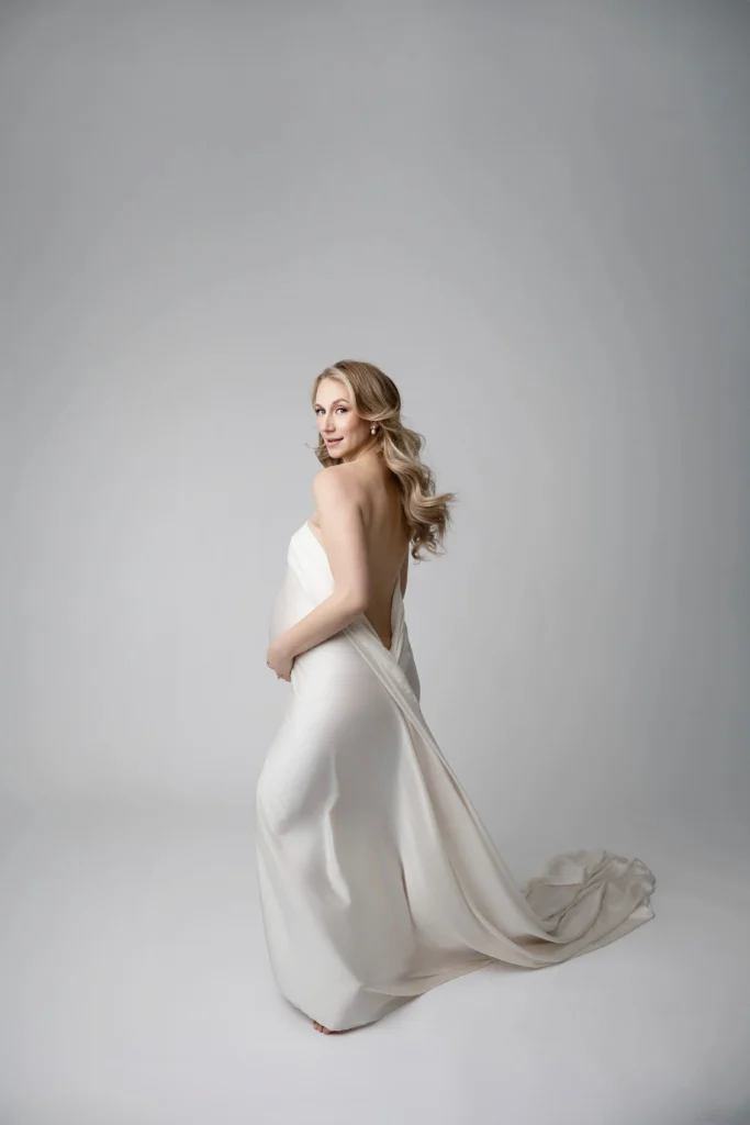 Woman in a white off the shoulder gown posing with her back to the camera in a studio satin fabric trailing on the floor