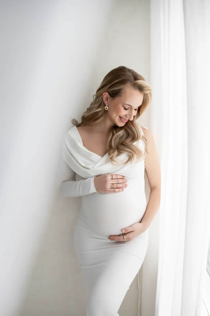 Pregnant woman in a white dress gently cradling her belly beside a bright window with sheer curtains