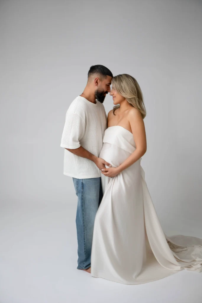 Couple facing each other in a tender moment pregnant woman in a white strapless dress cradles her belly with her partner beside her in a white knit top and jeans