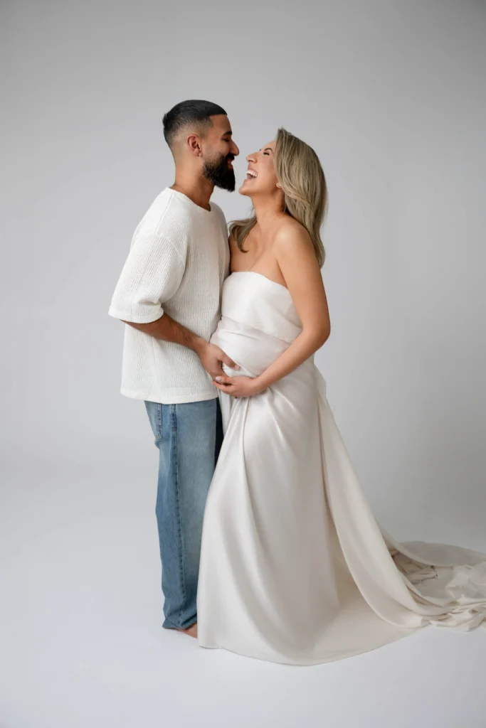 Couple standing close together the pregnant woman in a white dress laughing as the man in a cream knit sweater gently touches her belly