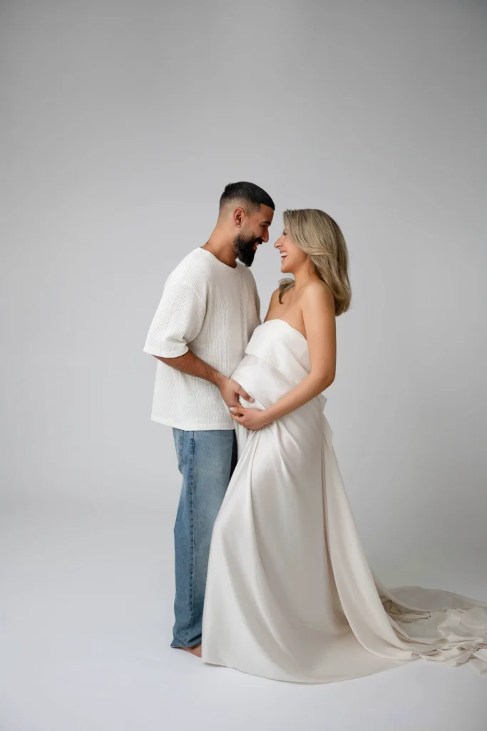 Happy couple standing close together smiling at each other the pregnant woman in a white dress holds her baby bump while the man in a cream sweater stands beside her