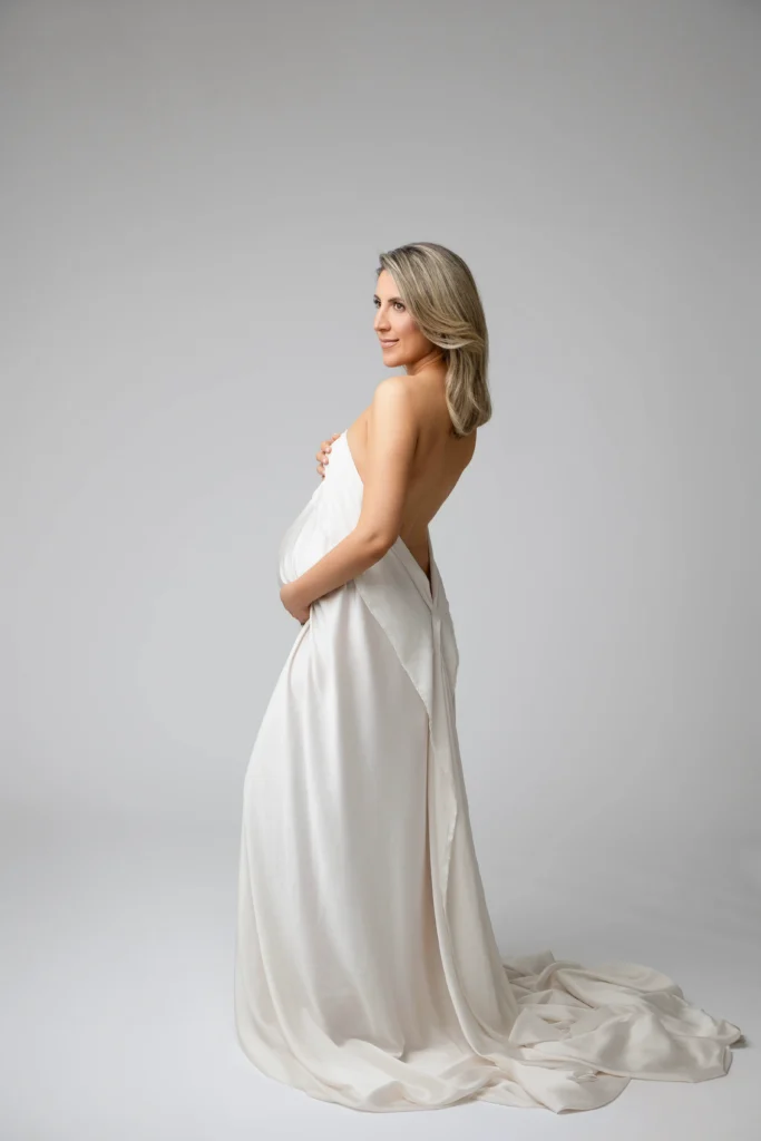 Pregnant woman standing in a flowing white fabric dress turned to the side hands resting on her baby bump against a neutral gray background