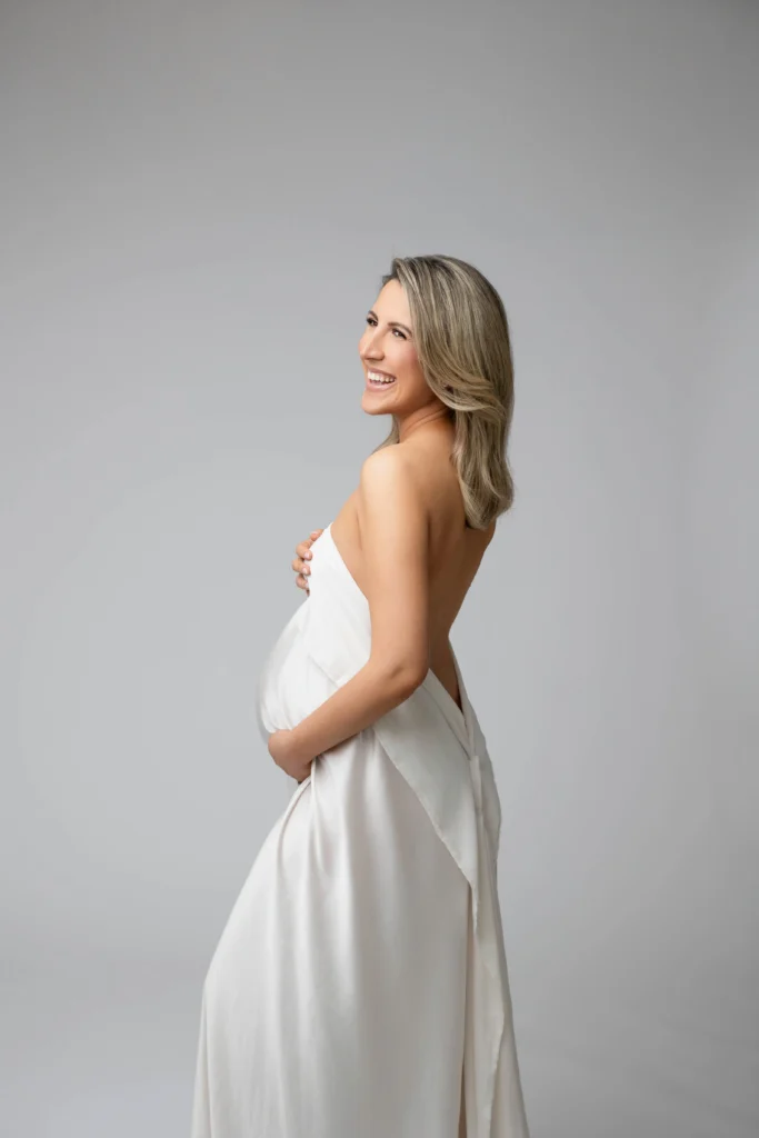 Side view of a pregnant woman in a white dress smiling and cradling her belly against a gray studio background