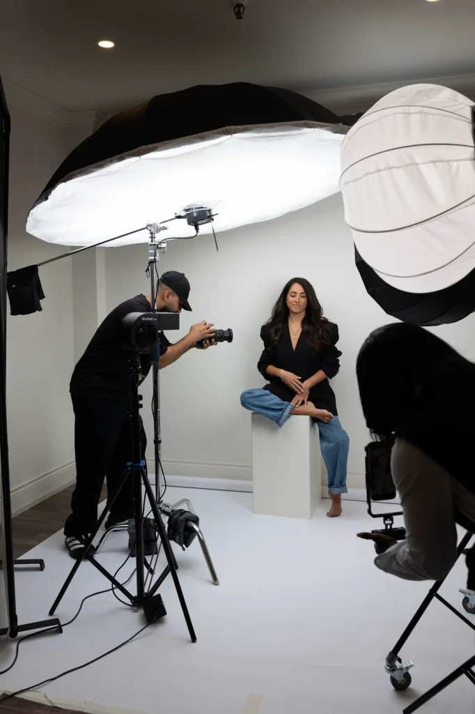 Photographer shooting a woman seated on a white pedestal in a studio with oversized softbox lights overhead